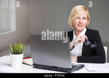Hübsche Blondine Geschäftsfrau im schwarzen Anzug am Arbeitsplatz Tisch sitzen mit Laptop in Weiß gekleidet im Büro Stockfoto