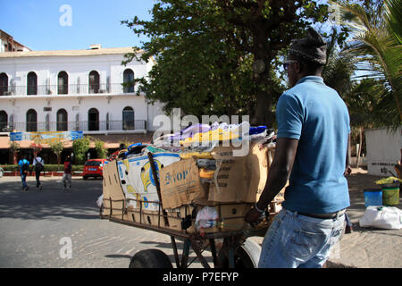 Afrikanischer Mann drückt seine Karre voller Schuhe und Sandalen auf den Markt von Saint-Louis-du-Senegal (Hotel de la Poste im Hintergrund) Stockfoto