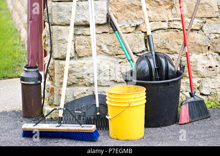 Reinigung von Werkzeugen gegen die Wand gelehnt. Ausrüstung für outdoor Reinigung. Besen, Rechen, Kunststoff gelb und schwarzen Eimer, Schaufel aus schwarzem Kunststoff.