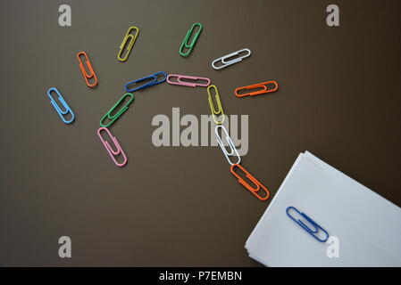 Farbige Büroklammern legte mit einem Fragezeichen und mit Blatt weißes Papier für die Aufzeichnung auf einem braun matt Hintergrund. Stockfoto
