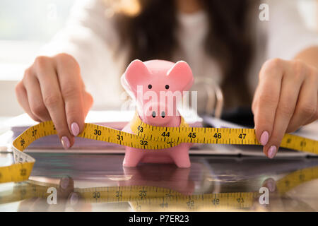 Die Geschäftsfrau Hand messen Sparschwein mit gelbem Maßband auf Glas Schreibtisch Stockfoto