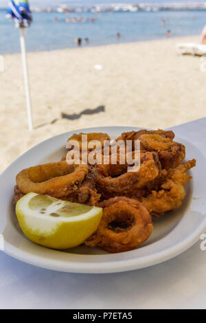 Griechische Taverne essen im Restaurant. Frittierte Tintenfischringe in Scheiben auf einem weißen Teller serviert mit Zitrone in unscharfer Strand Hintergrund. Stockfoto