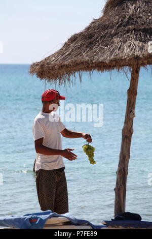 Tunesischen Anbieter in Red Hat, weißes T-Shirt bietet Trauben zu Touristen auf Mediterranean Beach Hotel in Hammamet in der Nähe von Meer im Sommer am Nachmittag im August Stockfoto