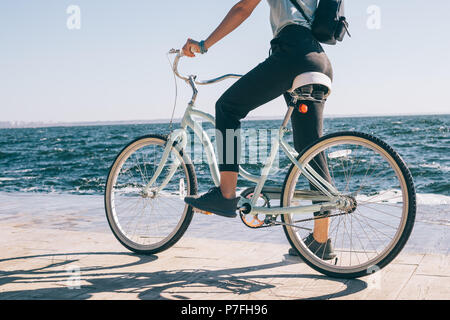 Rückansicht junge Frau während ein Fahrrad das Meer an sonnigen Sommertagen Fahrt gestoppt. Frauen tragen Rucksack stehend einen Fuß auf Pedal von Cruiser Fahrrad Stockfoto