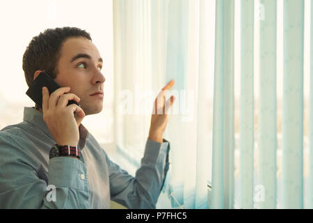 Executive über sein Mobiltelefon. Der Kerl im blauen Hemd ist, am Telefon zu sprechen vor dem Fenster und öffnet die vertikale Jalousien. Vertikale Stockfoto