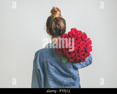 Zurück Blick auf die junge Frau in Jeans Jacke holding Strauß roter Rosen auf Schulter. Mädchen mit bun Hochsteckfrisur in Jeans mit Blumen. Weißer Hintergrund. Kopieren Sie sp Stockfoto