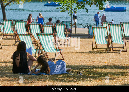 Zwei junge Frauen, die in der Sonne im Hyde Park, London UK sitzen, während der Juli 2018 Hitzewelle, mit Liegestühlen und Boote auf dem See, im Hintergrund Stockfoto