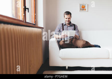 Schöner Mann auf dem Sofa zu Hause sitzen, mit einem digitalen Tablet Stockfoto