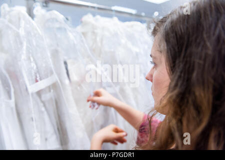 Glückliche junge Frau suchen, die versuchen, auf, die Auswahl Brautkleider in Boutique Discounter, viele weiße Kleider hängen an Rack Kleiderbügel Stockfoto