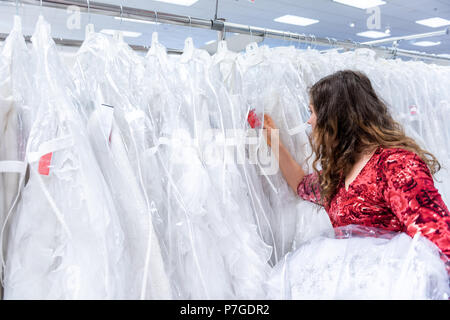 Junge Frau sucht und versucht, auf, Browsen, berühren, Kontrolle der Wedding Dress in Boutique Discounter, viele weiße Kleider hängen am Rack Stockfoto