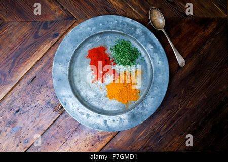 Ein Stilleben Fotografie eines rustikalen Rundschreiben silber Teller mit drei bunten gehackte Gewürze, roter Chili, orange Gelbwurz und Grünen corriander, auf einer Stockfoto