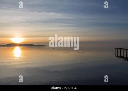 Ein Shooting für ein Sonnenuntergang über einem See, mit Sonne, die hinter einer Insel und ein Pier auf der rechten Seite Stockfoto