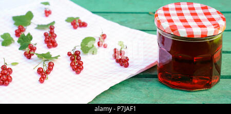 Rote Johannisbeeren liegen in Trauben auf karierte Tischdecke. Daneben ist ein Glas roten Johannisbeeren Gelee. Grünes Holz Hintergrund. Stockfoto