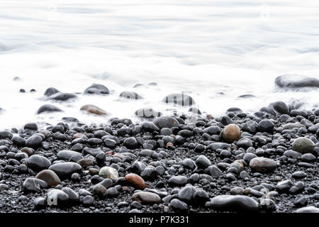 Nahaufnahme des großen schwarzen vulkanischen Felsen auf Sand Strand Reynisfjara, Island, mit langen Belichtungszeiten glatte Wasser Wellen am Ufer, glänzende nasse Steine Stockfoto