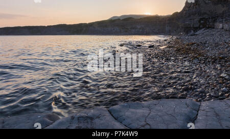 Kimmeridge bay Klippen in Dorset, Großbritannien ab dem Meeresspiegel während einer ruhigen Sommer Sonnenuntergang mit kleinen Wellen und die Sonne gesehen. Stockfoto