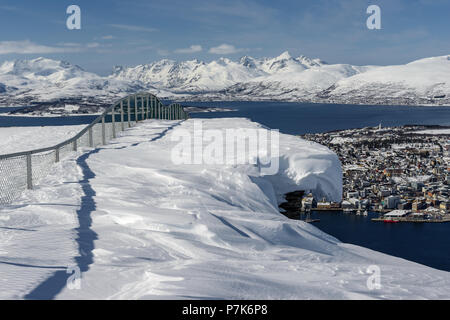 Schnee hängen über den Rand der Klippe. Tromsø und die umliegende Landschaft gesehen gleich außerhalb des Fjellstua (421 m). Fløya, Tromsø, Troms, Norwegen. Stockfoto