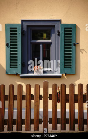 Ein Wohnhaus mit nostalgischen Fensterläden aus Holz, Fenster, Blumen und einem holzzaun vor dem Haus. Stockfoto