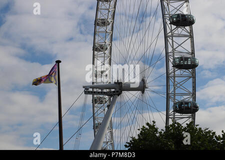 Der Blick auf das London Eye Riesenrad vor einem blauen Himmel mit weißen Wolken, und eine Fahne im Wind. Kapseln, gespannte Stahlseile, Achse. Stockfoto