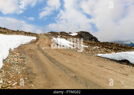 Dirt Road, steinigen Weg an die Spitze eines felsigen Berg bedeckt mit Schneeverwehungen, einem nebligen blauen Himmel im Hintergrund mit weißen Wolken im Sommer Stockfoto