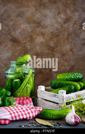 Pickles. Leckere Salzgurken in einem Glas, frische Ernte in einer Holzkiste, Gewürze, Kräuter auf einem Tisch. Vorbereitung von Gurken für Pickles. Vertikale Stockfoto