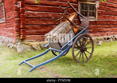 Ein blaues Holz- Vintage zwei Rädern Handwagen mit Stühlen und einem Trunk vor einem roten Holzhaus geladen. Stockfoto