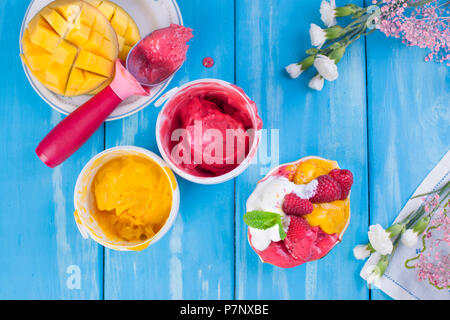 Eis in verschiedenen Farben auf blauem Hintergrund. Himbeeren und Mango. Einrichtung von Blumen Stockfoto