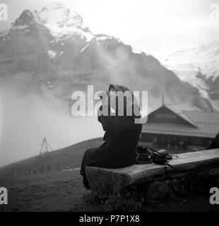 Eine ältere Frau sitzt in die Berge auf einer Holzbank und untersucht die Landschaft durch ein Fernglas, Ca. 1945 bis 1955 Stockfoto