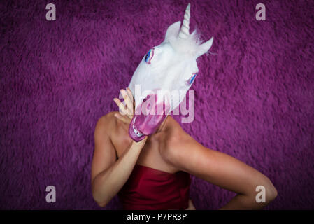 Funny Girl Einhorn flirte mit Ihnen. Freaky junge Frau in komischen Maske steht auf lila Hintergrund. Portrait von ungewöhnlichen Dame im roten Kleid Stockfoto