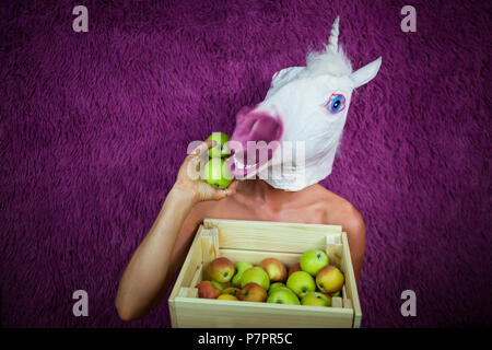 Freaky junge Frau in komischen Maske steht auf lila Hintergrund mit Box von Früchten. Porträt einer ungewöhnlichen Frau. Funny Girl Einhorn hält Äpfel in die Hand. Stockfoto