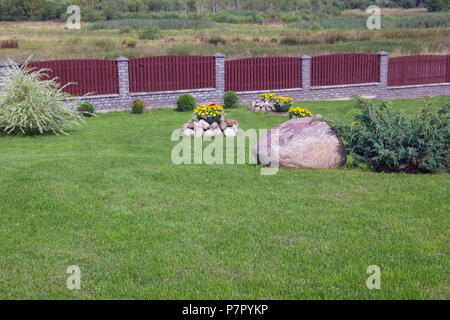 Teil der Grünen ländlichen Hof. Große Blumentöpfe mit gelben Blumen und dekorative Bush im Sommer rasen. Langen hölzernen Zaun schützt das Dorf Perimeter. Stockfoto