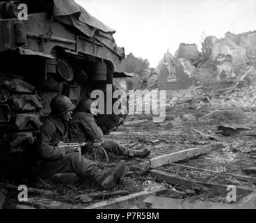 36Th AIR 9 ID Geich Dez 1944. Mit deutschen Schalen schreien Overhead, Amerikanische Infanteristen suchen Schutz hinter einem Panzer. Im Hintergrund können die Ruinen der Stadt Geich, Deutschland, die immer noch unter schweren Beschuss ist gesehen werden. Dezember 11, 1944. Roberts. (Armee) Stockfoto