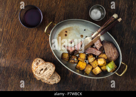 Rindersteak mit gebratenen Kürbis in der Pfanne auf dunklem Hintergrund mit einem Glas Rotwein. Tisch Konzept, Ansicht von oben Stockfoto