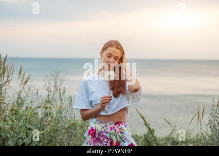 Gerne schöne Frau bläst Löwenzahn über Himmel und Meer Hintergrund, Spaß und Spielen im Freien, Natur, Sommer Urlaub und Ferien, Yo Stockfoto