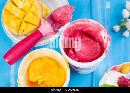 Fruchteis mit Mango und Himbeeren in einen Eimer packen. Löffel für Speiseeis. Blauer Hintergrund und Blumen. kopieren. Stockfoto