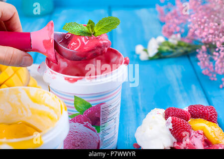 Fruchteis mit Mango und Himbeeren in einen Eimer packen. Löffel für Eis in der Hand. Blauer Hintergrund und Blumen. Karte Stockfoto