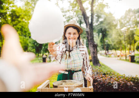 Foto Thema Small Business kochen Süßigkeiten. Eine Junge kaukasier Frau mit einer Schürze Trader in den Hut der Eigentümer der Steckdose macht eine Zuckerwatte, einer Messe Stockfoto