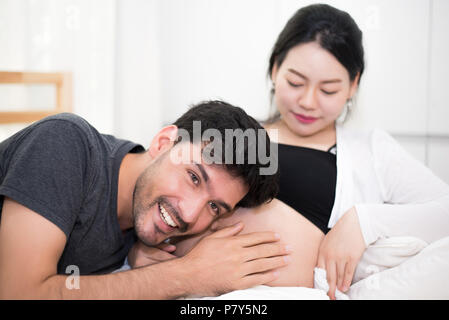Anhörung der Vater seinen Sohn oder seine Tochter kicking Sound innerhalb von Mutter beim Sitzen auf Bauch liegend auf Bett zu Hause. Familie und Freunde Konzept. Happy sweet home Stockfoto