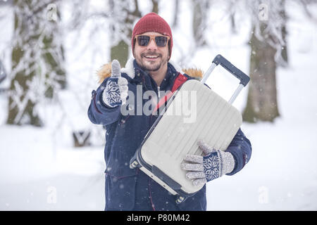 Porträt der jungen modische Mann auf Schnee holding Koffer und Daumen hoch. Winter Ferienhäuser Travel Concept. Stockfoto