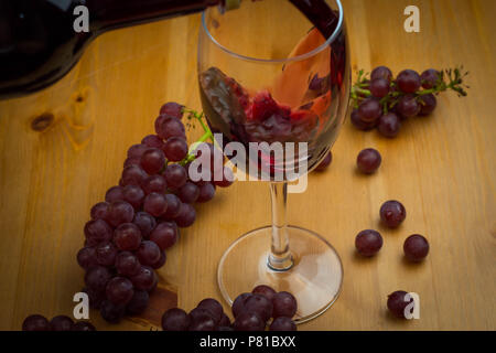 Rotwein gegossen in ein Glas Wein auf einen hölzernen Tisch mit frischen Weintrauben als Hintergrund design Stockfoto