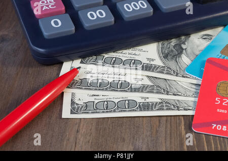 Banknoten für hundert Dollar, Kreditkarten, einen Taschenrechner und einen Stift auf einem Holztisch. Stockfoto