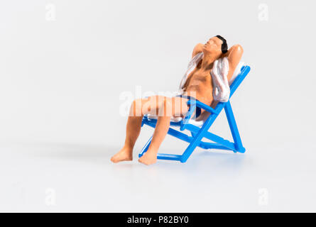 Miniatur Mann in T-Shirt und Badehose entspannenden Sonnenbad auf einer klappbaren blau Liegestuhl Sessel mit seiner Hand in den Nacken konzeptionelle eines Sommers Stockfoto