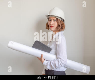 Schöne asiatische Frau Ingenieur curly kurze Haare tragen weiße Schutzhelm und weißes Hemd holding Blueprint und Notebook auf die weiße Wand im Hintergrund. W Stockfoto