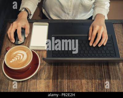 Hand tippen auf schwarzer Laptop Computer und klicken Sie auf Computer Maus, bei Arbeiten mit Kaffee und Handy auf hölzernen Tischplatte in caf konzentriert Stockfoto