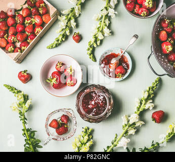 Flach frische Erdbeeren in Holz- Sieb und Schüsseln mit Jam jar und Löffel auf dem Küchentisch Hintergrund mit Garten Blumen, Ansicht von oben. Sommer berr Stockfoto