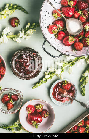 Frische Erdbeeren in Sieb und Schüsseln mit Jam jar und Löffel auf dem Küchentisch Hintergrund mit Garten Blumen, Ansicht von oben. Sommer Beeren con bewahren Stockfoto