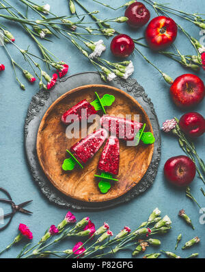 Holzplatte mit hausgemachten rote Früchte Eis oder Popsicle gefrorenes Obst Saft auf rustikale Küche Tisch Hintergrund mit Garten Blumen und Zutaten, Stockfoto