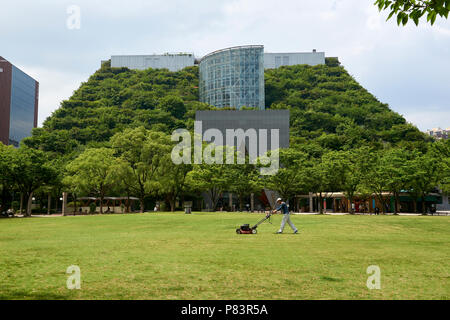 Ein Mann mäht den Rasen in Tenjin Central Park vor der Umweltfreundliche ACROS Gebäude in Fukuoka, Japan. Stockfoto