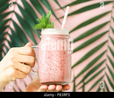 Fands Holding eine Frucht pink Smoothie in einem Glas gegen eine dekorierte rosa Wand mit einem Palm Leaf. Im minimalistischen Stil. Trend vintage getönt. Stockfoto