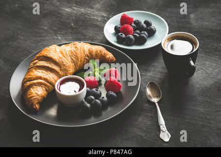 Französische Croissant, Beeren, Marmelade und Kaffee auf Schwarzen Tisch, kontinentales Frühstück. Getonten Bild Stockfoto
