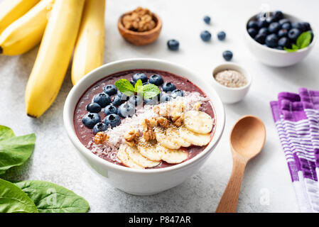 Acai smoothie Schüssel mit superfood Belag auf konkreten Hintergrund. Lila Smoothie in Weiß Schüssel mit Banane, Chia Samen, Kokos, Walnüsse und blueber Stockfoto